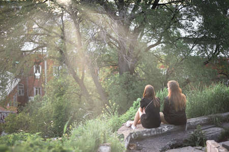 Two young women are sitting on a hillside admiring nature and chatting in the rays of the sun at sunsetの写真素材