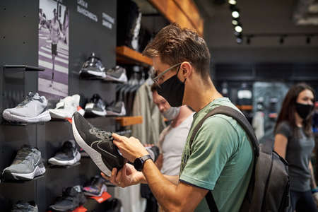 Nizniy Novgorod / Russia - 15 07 2020: Young Man with medical face mask chooses sneakers in shopping store. New reality after covid 19 outbreak.のeditorial素材