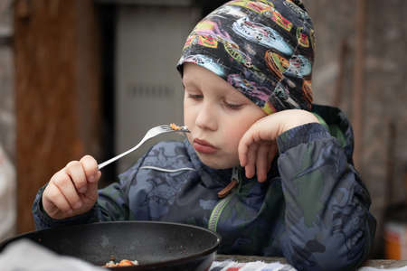 Close up of cute thoughtful preschool child eating by himself. Hungry boy eating his lunch by himself and sitting alone at table outside. Independence of childのeditorial素材