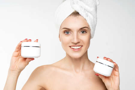 smiling girl holds in her hand two jars with cosmetic, a towel on his head