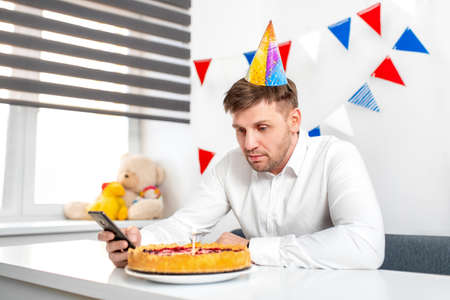 Upset young man party cap sitting at the birthday cake and looking with sad eyes on it. Concept of celebrating alone. Quarantine.の写真素材