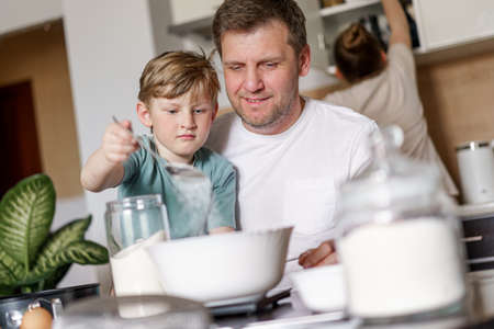 Dad and little son baking together in kitchen, preparing dough for pie, having fun at home during...