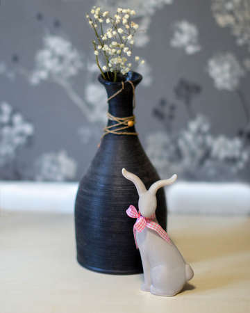 A small, beautiful gray ceramic rabbit with a red checkered bow on the table next to the vase.の写真素材