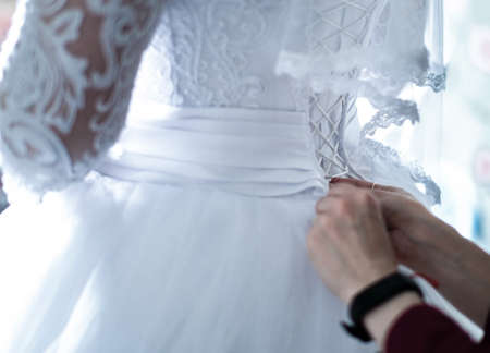 Bridesmaid ties the bride corset. Lace long veil. Bride white dress. Laces on the corset of the...