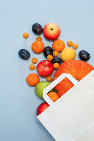 Package with scattered colored fruit on a blue background. Plums, tangerines, lemon, apples, pumpkin. Shopping in the store. Vertical arrangementの写真素材