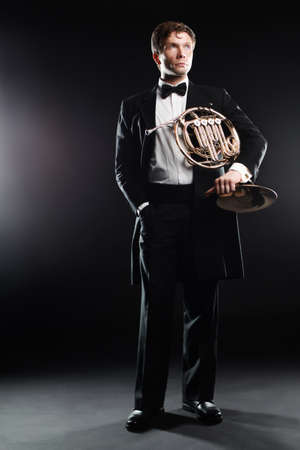 Elegant man in tuxedo with bow tie. Classical fashion man musician with french horn classic instrument. Full length portrait of hornist music playerの写真素材