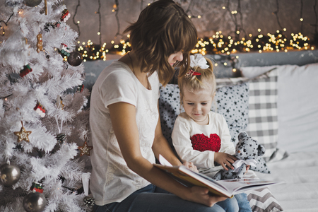 Little girl with mother reading a book sitting under the Christmas tree.の写真素材