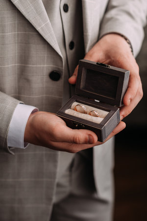 The image shows a man indoors wearing a suit and tie, holding a wooden box with two rings inside.