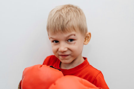 Boy with a scratch on his face giving a glare at the camera, wearing boxing gloves on his hands, set against a white backgroundの写真素材