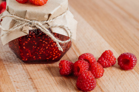 Raspberry jam in a jar on a wooden board, the lid is decorated with paper and tied with twine....