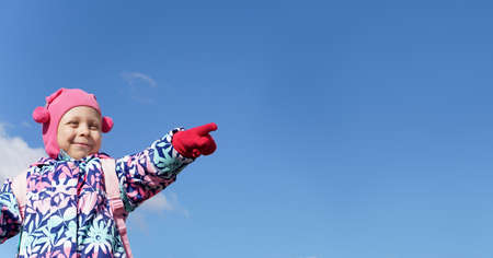 A smiling girl in a pink hat and gloves shows the direction. Child on the background of the blue sky. The cold season is autumn, winter, spring. Concept for bannerの写真素材