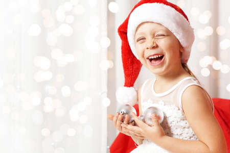 Cheerful little girl in a santa claus cap laughs, bokeh in the background. Lights, glowing background, blurred background. Place for your text. Card. Congratulation. The symbol of the new year. 2022の写真素材