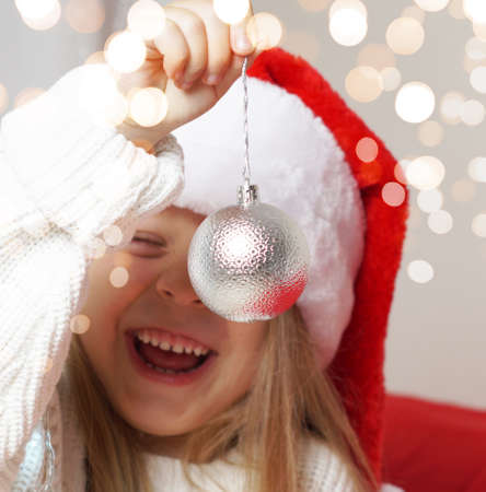 A Christmas decoration is large in the foreground. In the background, a blurry little girl in a santa hat. Silver ball on the Christmas tree. 2022. New Years Concept. Golden bokehの写真素材