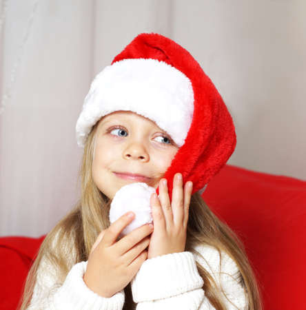 A cute girl in a red cap holds a pompom. The child wishes a Happy Christmas and New Year. Card. Congratulation. The symbol of the new year. 2022の写真素材