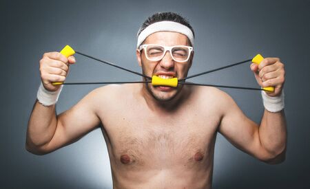 Silly aggressive man exercising over dark gray background. Nerd guy with eyeglasses holding expander. Close up of funny young man  doing aerobics. Studio shotの写真素材