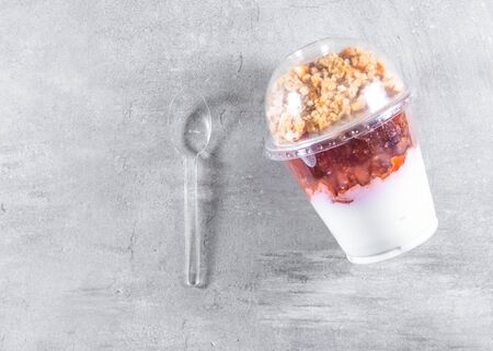 Yogurt with granola flakes and fruits in a plastic cup to go over gray concrete background. Flat layの写真素材