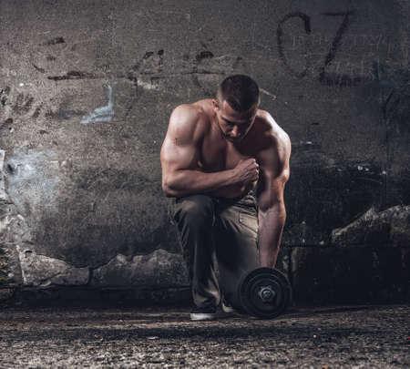 Bodybuilder with weight, strong athlete holding dumbell against grunge wall, dramatic light, dark indoors, copyspace for textの写真素材
