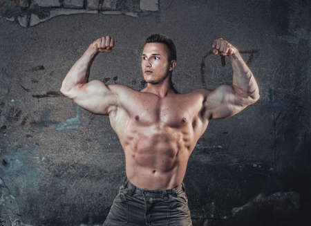 Bodybuilder showing hic biceps, young muscular man against damaged old wall