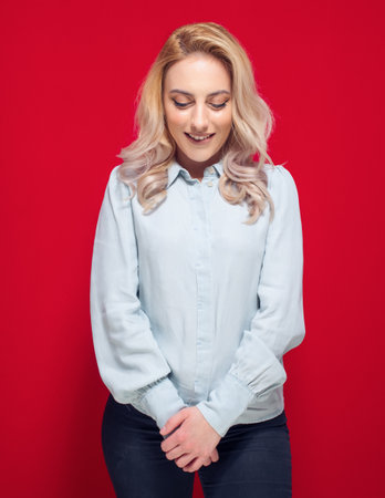 Lovely girl with closed eyes and smiling face looking down, isolated on red background. Young woman with hand on hand. Happiness concept. Studio shotの写真素材