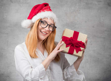 Young beautiful blond woman wears christmas santa claus hat - cap and holding red gift box - present with ribbon, isolated on gray background. Studio shotの写真素材