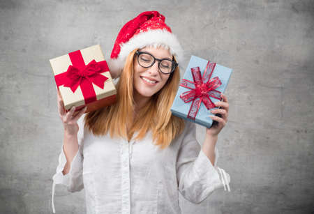 Santa woman holding christmas gifts isolated on gray background. Happy young girl wearing red santa hat and holding present box. Studio shotの写真素材