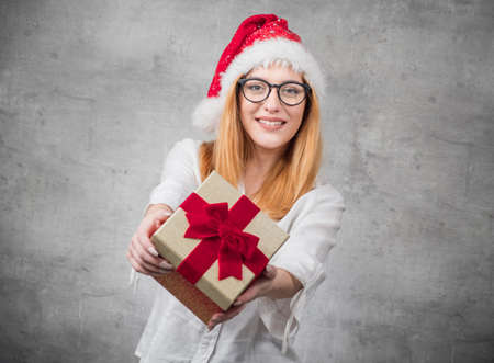 Santa woman giving christmas present box isolated on gray background. FOCUS ON BOX. Happy young girl holding gift. Studio shotの写真素材