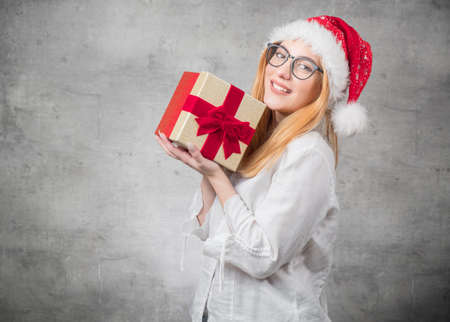 Happy beautiful blond woman wears christmas santa claus hat - cap and holding red gift box - present with ribbon, isolated on gray background. Studio shotの写真素材