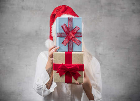 Santa woman holding christmas gifts and covering his face, isolated on gray background. Happy young girl wearing red santa hat and holding present box. Studio shotの写真素材