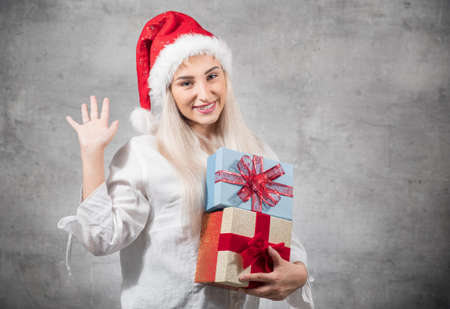 Playful santa woman holding christmas gifts isolated on gray background. Happy young girl wearing red santa hat and holding present box. Studio shotの写真素材