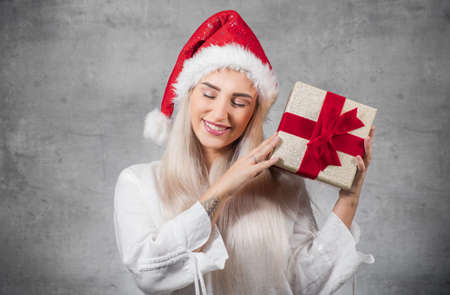 Lovely woman wears christmas santa claus hat - cap and holding red gift box - present with ribbon, isolated on gray background. Studio shotの写真素材