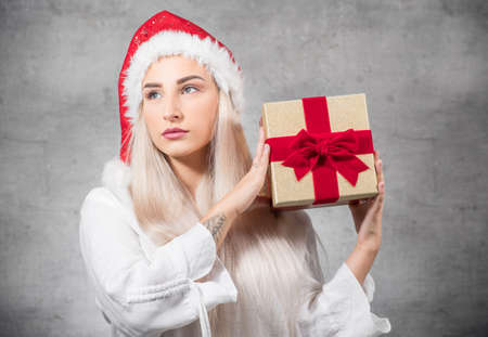 Santa woman with christmas gift isolated on gray background. Thinkful young girl wearing red santa hat and holding present box. Studio shotの写真素材