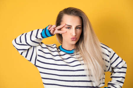 Amusing girl shows peace sing with fingers and send kiss to you, isolated on yellow background. Joyous lovely woman kissing. Studio shotの写真素材