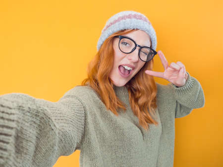 Trendy girl holding in hands smartphone and making selfie while showing v-sign, isolated on yellow background. Young woman using smart phone. Studio shotの写真素材