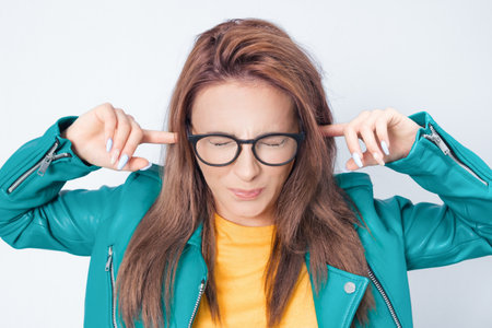 Noise - loud. Closeup portrait of a young angry unhappy stressed woman covering his ears closing eyes, wearing green leather jacket, isolated on white background. Studio shotの写真素材