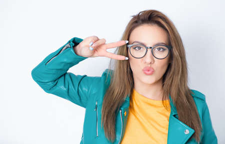 Amusing girl shows peace sing with fingers and send kiss to you, wearing green leather jacket, isolated on white background. Joyous lovely woman kissing. Studio shotの写真素材