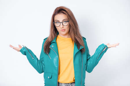 Clueless young woman against white background being in confusion with stretched arms opened palms, wearing green leather jacket, isolated on white background. Studio shotの写真素材