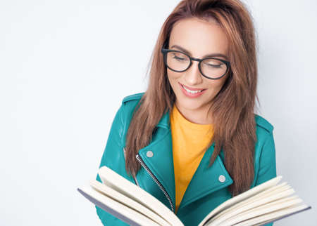 Happy young woman reading book while standing, wearing green leather jacket, isolated on white background. Cheerful cute girl with a book. Studio shotの写真素材