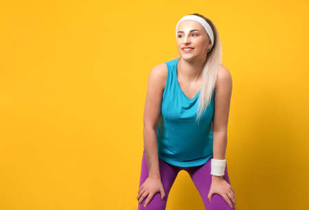 Fitness young woman wearing sportswear,sport clothing, posing and looking to side while keeps her arms on legs,leg, isolated on yellow background. Athlete female person. 80s styling. Studio shotの写真素材