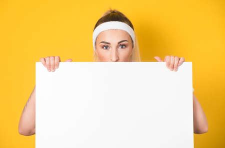 Surprised face of young women behind white board, isolated on yellow background. One fitness girl showing blank empty paper billboard with blank space near her head. 80s styling. Studio shotの写真素材