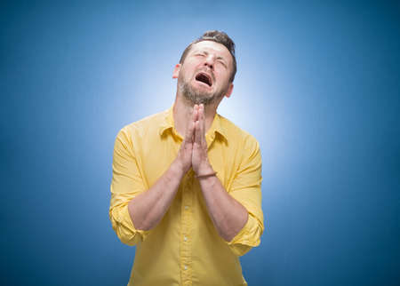 Crying disappointed young man asking for forgiveness over blue background, dresses in yellow shirt. Sad guy looking up. Guilt concept. Studio shotの写真素材