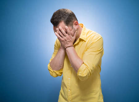 Forgetful man holding his hand on face over blue background, dresses in yellow shirt. Forget - forgot concept. Studio shotの写真素材