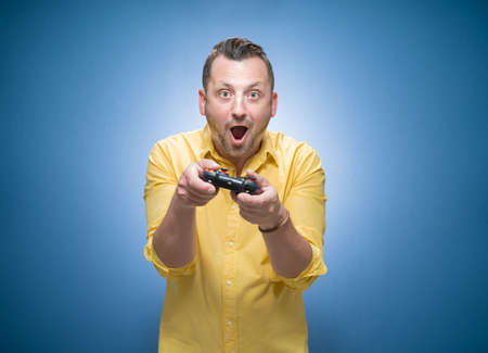 Surprised man playing video games with joystick over blue background, dresses in yellow shirt. Astonished guy holding game controller and looking at camera. Studio shotの写真素材