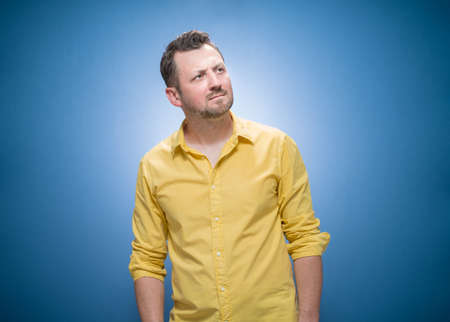 Strict young man thinks. Portrait of doubtful guy with confusion face expression over blue background, dresses in yellow shirt. Careful man, copy space for text. Studio shotの写真素材