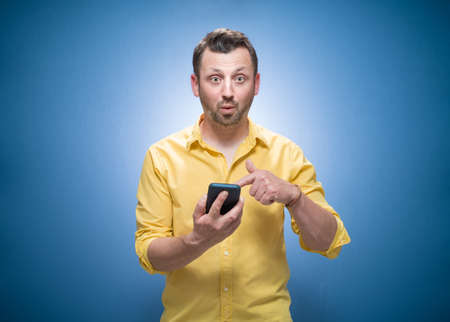Surprised young man shows the mobile phone blank display - space for your text or image over blue background, dresses in yellow shirt. Studio shotの写真素材