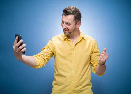 Young man holding in hands smartphone and making selfie while showing v-sign over blue background, dresses in yellow shirt. Trendy guy using smart phone. Studio shotの写真素材