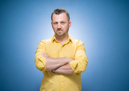 Portrait of serious young man, strict guy with hands on waist looking to camera over blue background, dresses in yellow shirt. Studio shotの写真素材