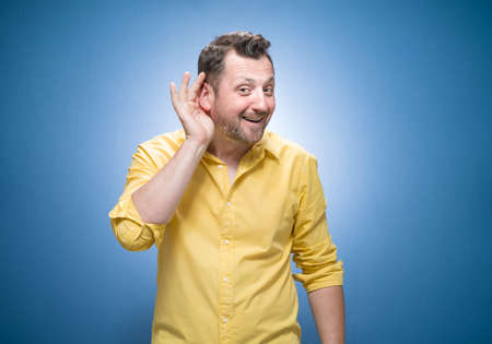 Half length man holding palm near his ear trying to listen something interesting over blue background, dresses in yellow shirt. Gossip concept. Studio shotの写真素材