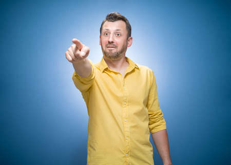 Prank man laughing at you and pointing with finger to the camera over blue background, dresses in yellow shirt. One guy with laugh expression. Prank and joke. Studio shotの写真素材