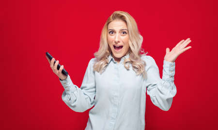 Trendy woman with phone unexpected incredible notification, isolated on red background. Amazed girl using smart phone . Studio shotの写真素材