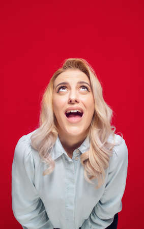Surprised young woman looking up to free space - copy space for your product or text, isolated on red background. Studio shotの写真素材